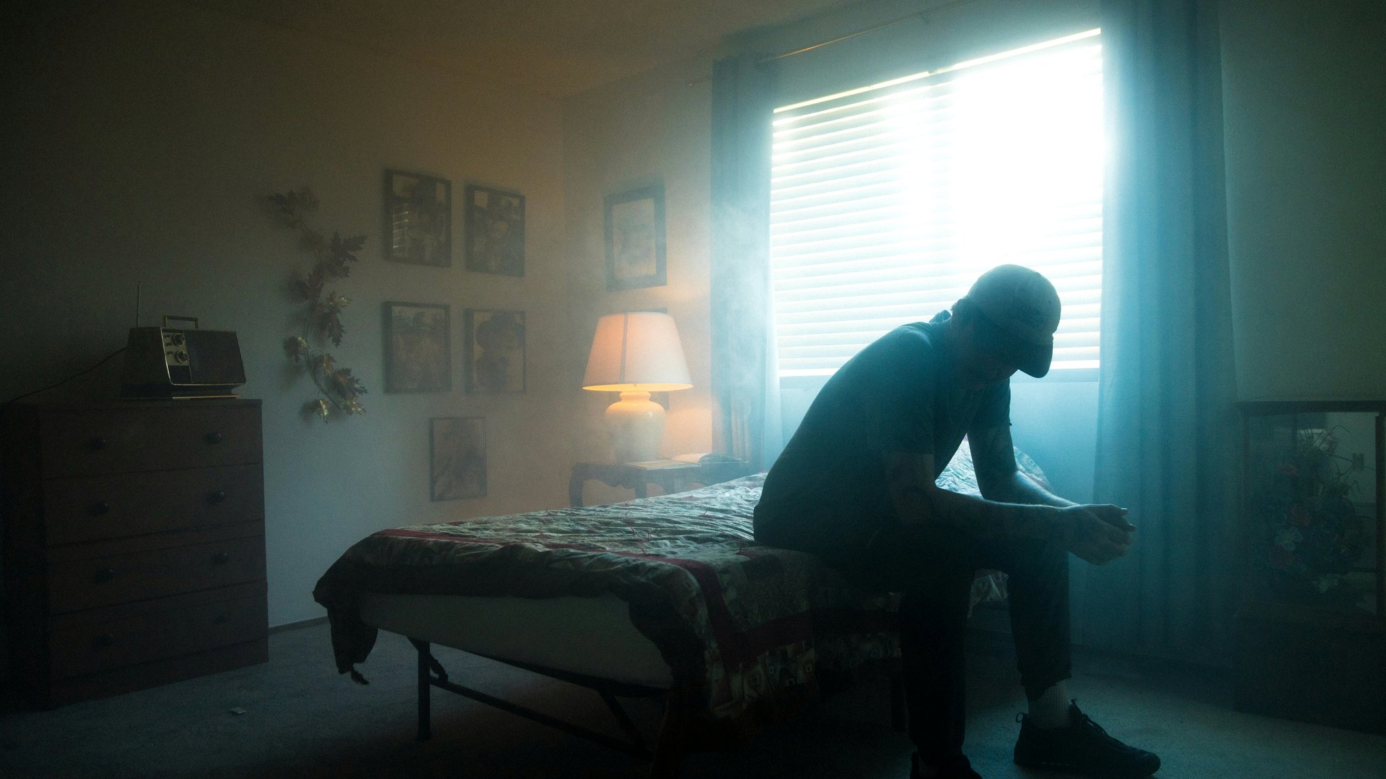 A man sitting on the edge of a bed, head down, backlit by a window.